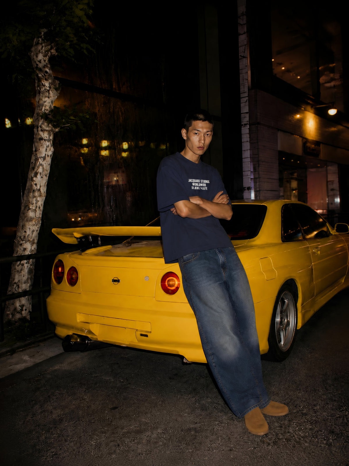 JacquardStudios Night Drive lookbook: model in a navy JacquardStudios Worldwide T-shirt leaning against a yellow car at night