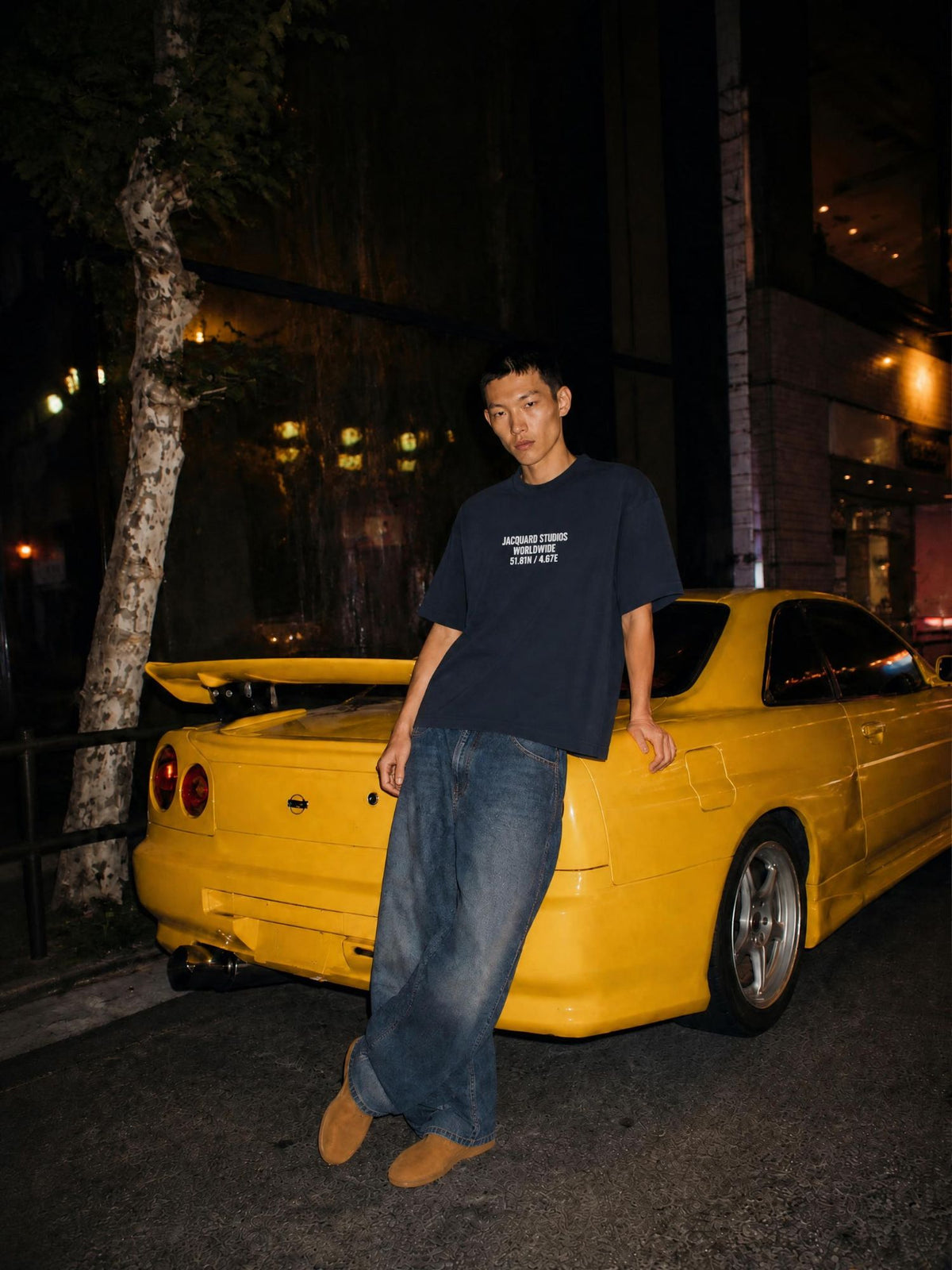 JacquardStudios Night Drive lookbook: model in the navy T-shirt and wide denim beside a yellow car on a night street